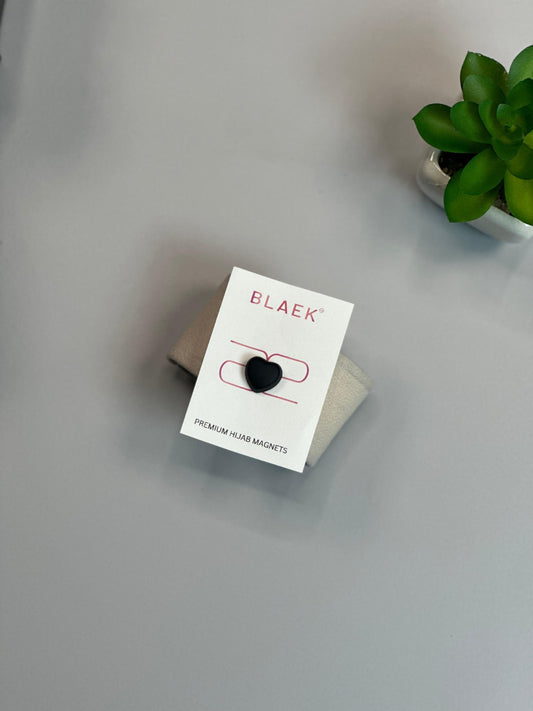 Black heart-shaped hijab magnet on premium packaging, placed on a neutral fabric.